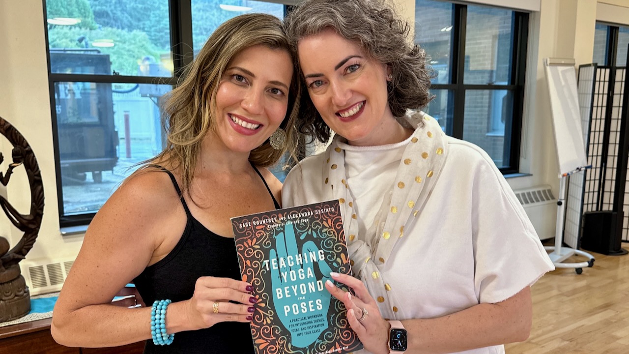 Alexandra and Sage, middle-aged smiling white women, pose with a copy of their book TEACHING YOGA BEYOND THE POSES