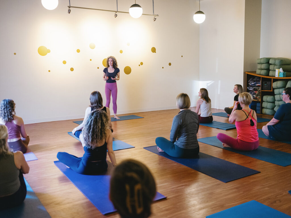 Sage Rountree talks to a class at Carrboro Yoga Company. She is standing; they are sitting.