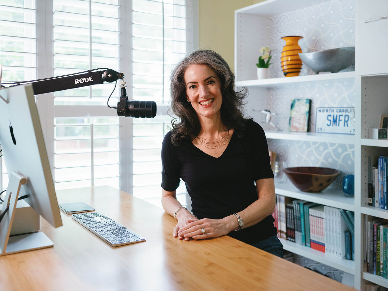 Sage Rountree, host of the Yoga Teacher Confidential podcast, sits at her desk with a microphone. She is a middle-aged white woman with graying hair in a black shirt, smiling.