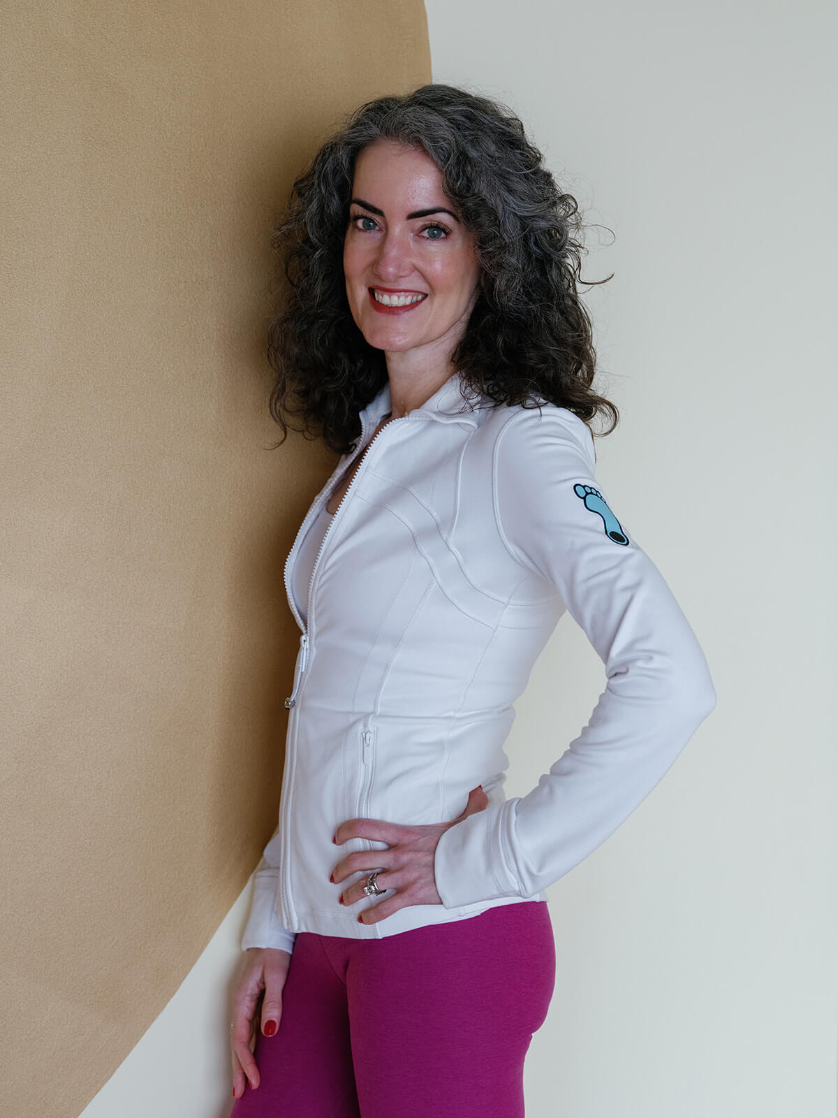 Carrboro Yoga Company Sage Rountree stands in her yoga studio wearing a jacket with a UNC Tar Heel. She is a middle-aged white woman with dark curly hair.
