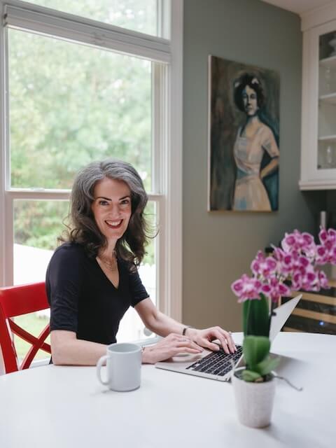 Sage Rountree sits with her laptop, smiling at the camera. She is a middle-aged white woman with graying hair and blue eyes.