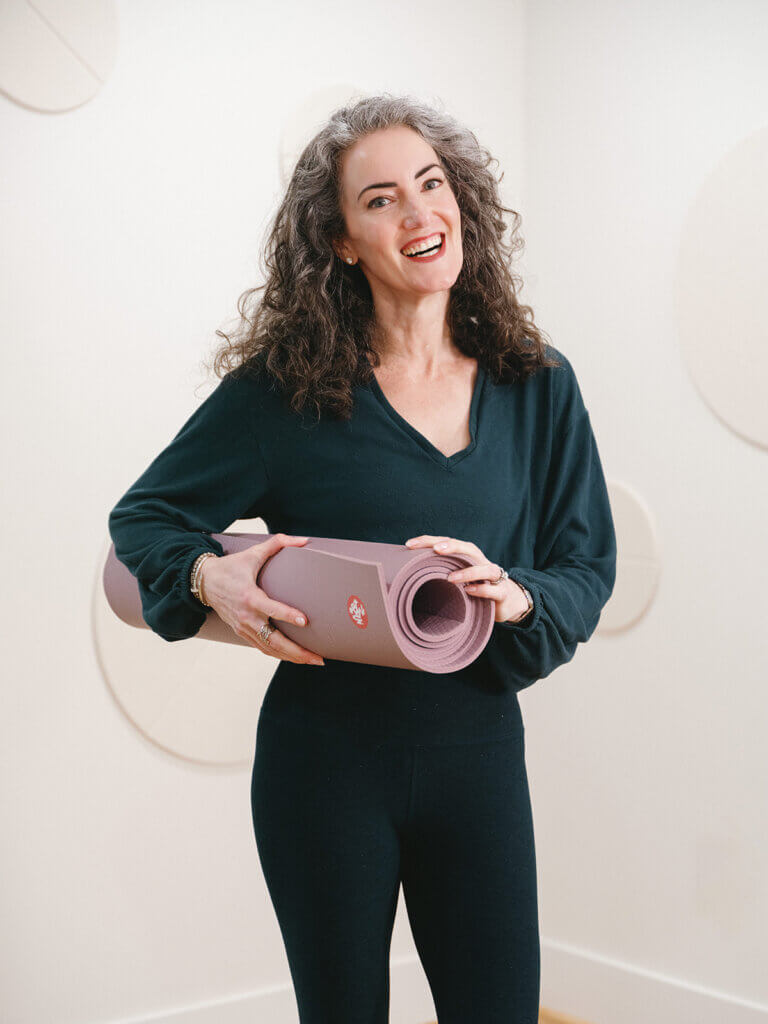 Sage Rountree smiles in her home studio, holding her yoga mat