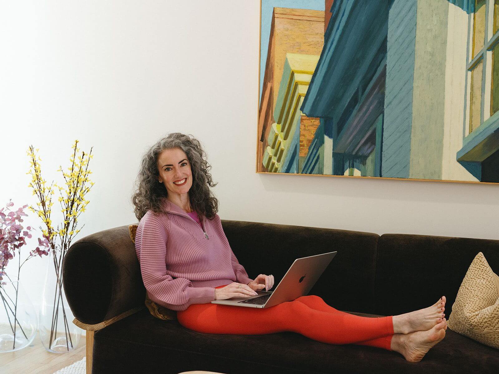 Sage Rountree smiles from a couch as she writes Sage Advice, her newsletter for yoga teachers, on her laptop