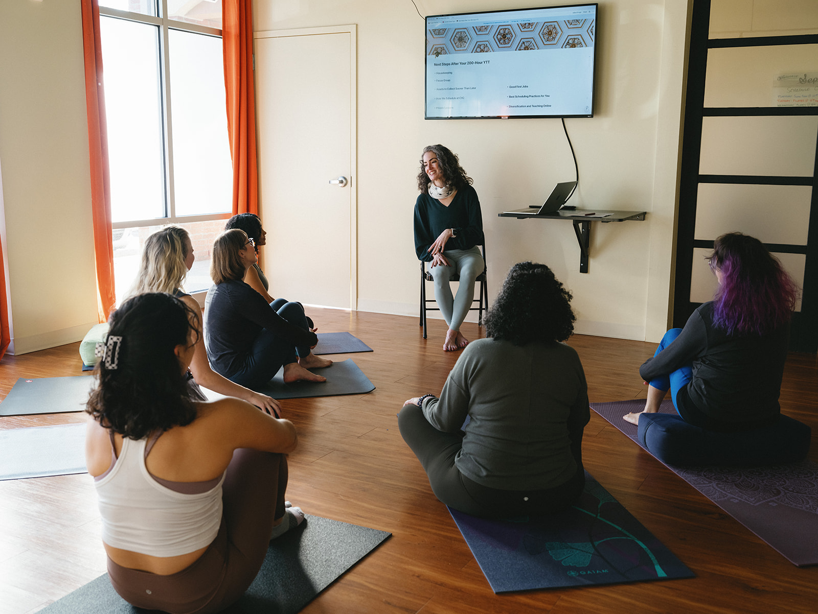 Questions to Ask Yoga Teacher Training Programs (and What Good Answers Sound Like) Sage Rountree sits on a stool beside a slide screen, teaching yoga teacher trainees seated on their mats in a sunlit studio
