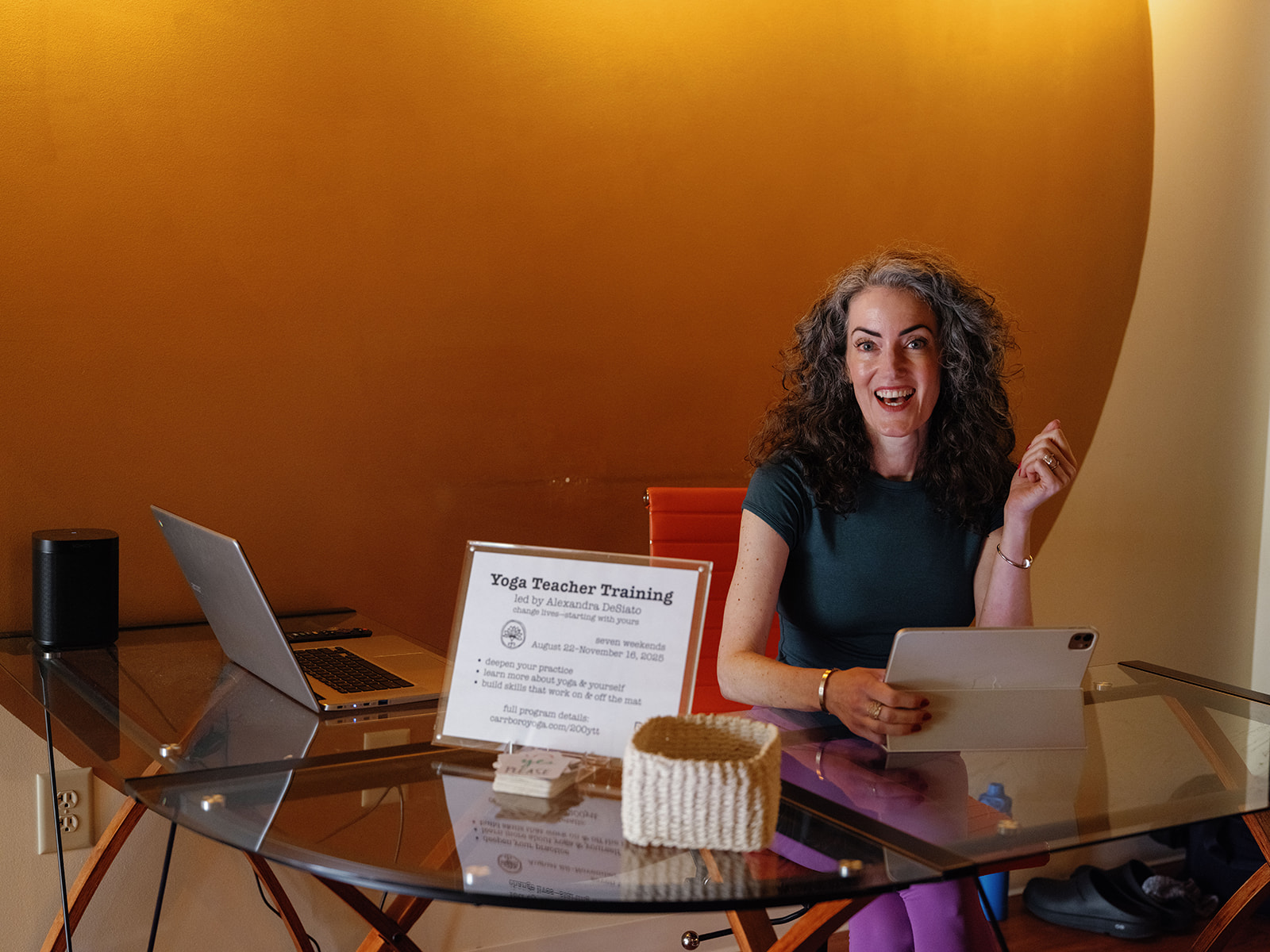 Yoga Alliance Changes for 2027: What’s Real, What’s Rumor Sage Rountree smiles at a glass desk beside a “Yoga Teacher Training” sign in a studio lobby