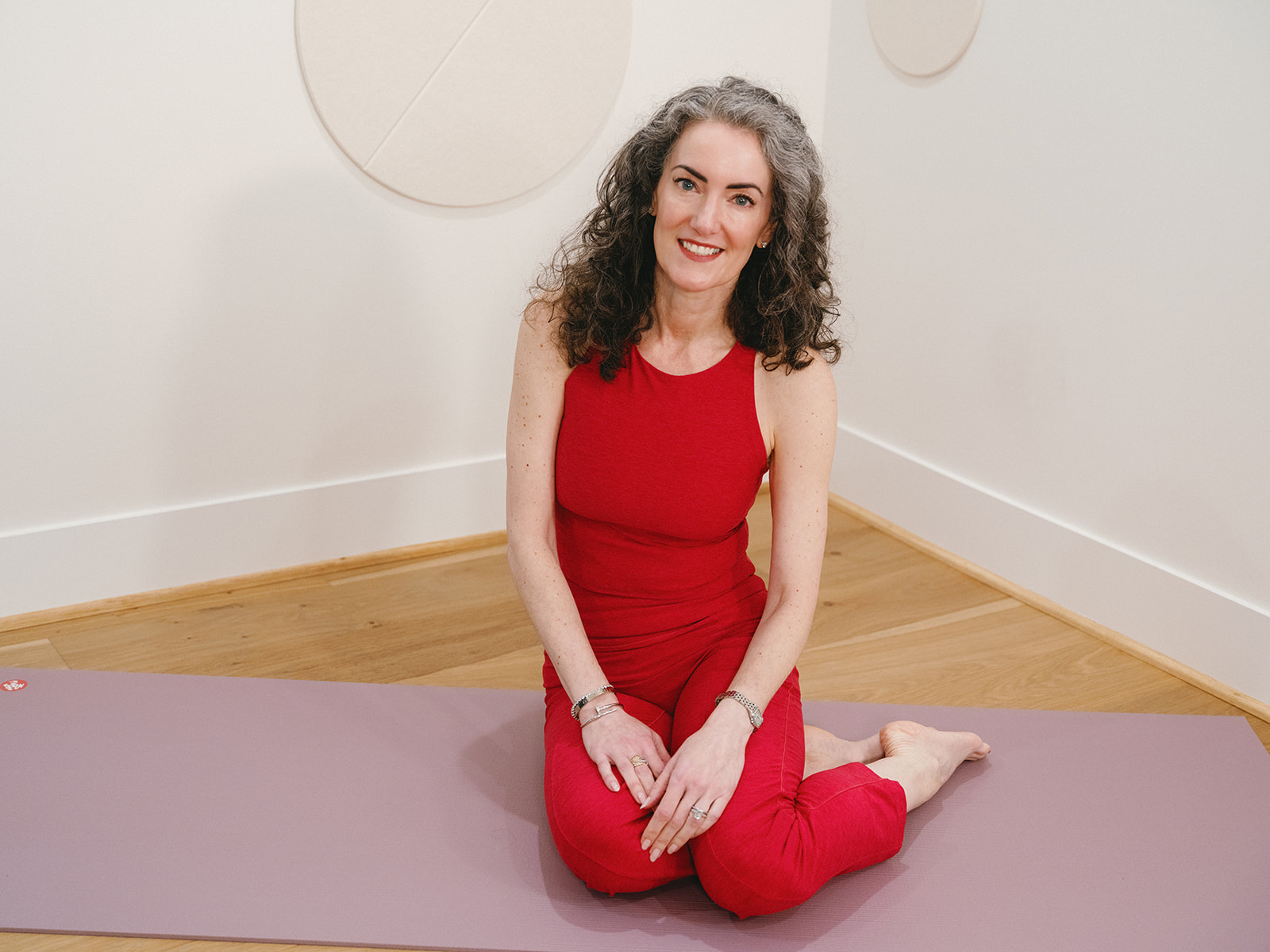 Sage Rountree, dressed in red, sits on a yoga mat in a bright studio and smiles at the camera